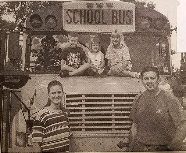 Mike and Jenny Aksamit standing next to one of their school buses with their three kids sitting on the hood of the bus